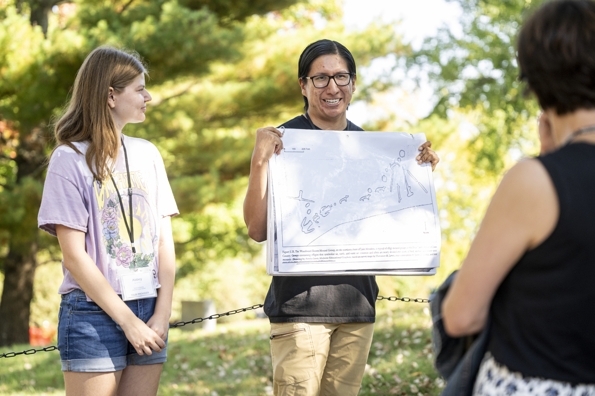 On a sunny day, two tour guides stand in front of a group. On the right, Kane Funmaker holds holds up a display graphic showing the location of a variety of burial mounds on the north shore of Lake Mendota.