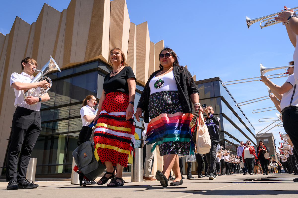 Two people in brightly patterned skirts walking with musicians playing instruments on both sides.