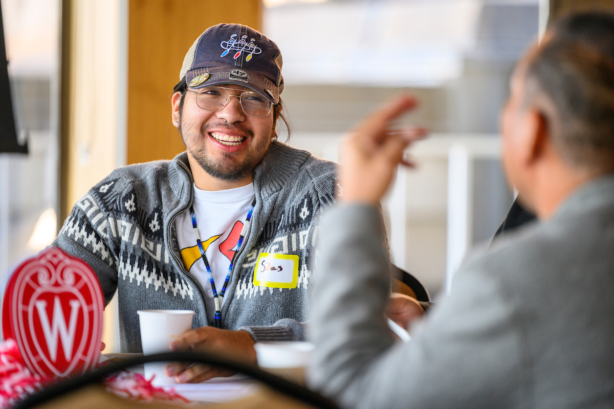 A person wearing a hat and glasses is smiling at someone across a table. The W crest is visible.