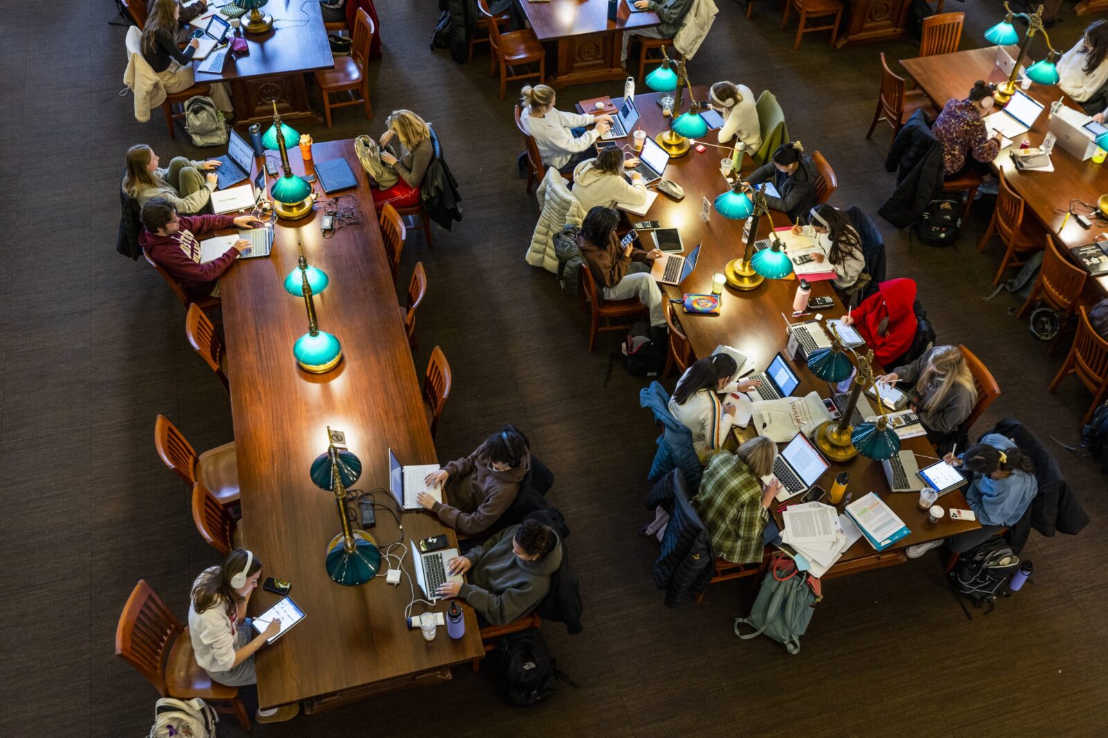 Many students are studying at long tables with green lamps. Photo taken from above.